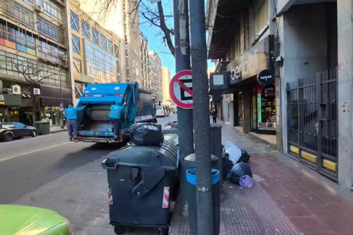 Camión de basura recogiendo residuos en una calle urbana con bolsas de basura acumuladas junto a un contenedor y un cartel de prohibido estacionar Camión de basura recogiendo residuos en una calle urbana con bolsas de basura acumuladas junto a un contenedor y un cartel de prohibido estacionar