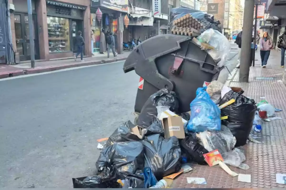 Contenedor de basura desbordado con bolsas y residuos acumulados en la vereda de una calle urbana Contenedor de basura desbordado con bolsas y residuos acumulados en la vereda de una calle urbana