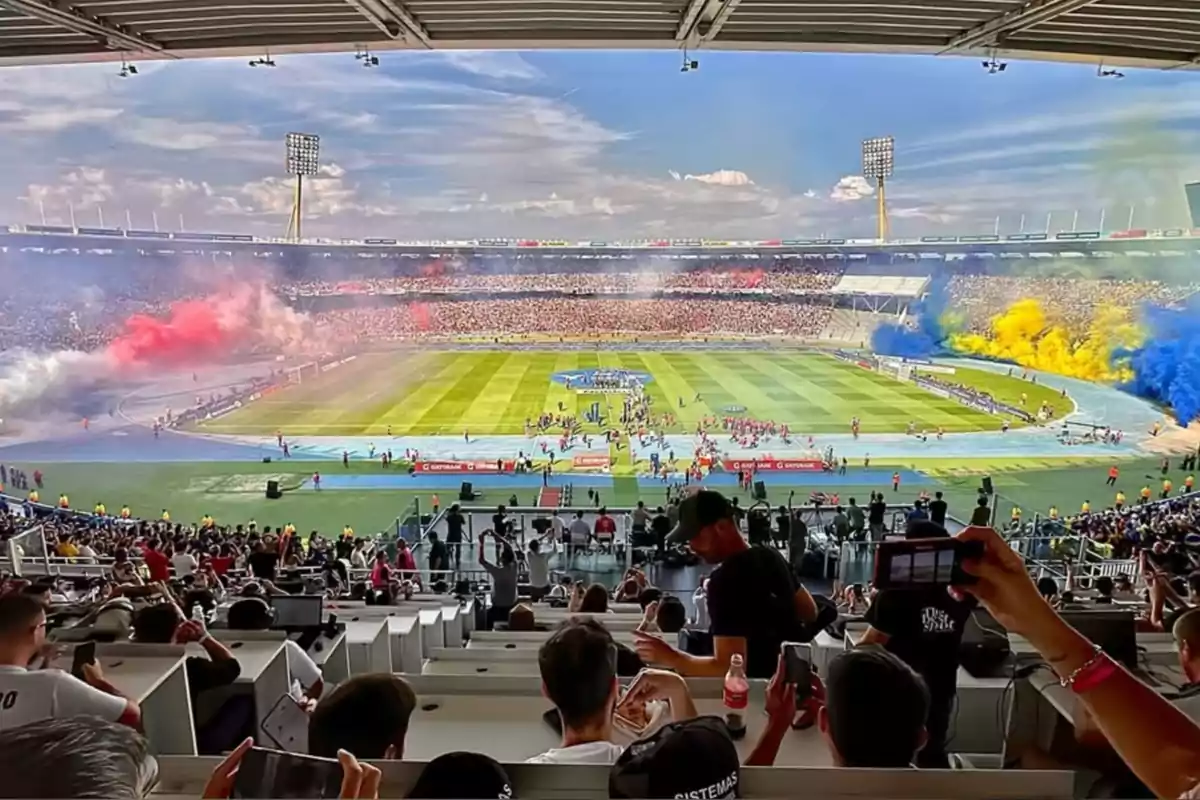 Estadio lleno de aficionados durante un partido de fútbol con humo de colores rojo, amarillo y azul en las gradas y personas tomando fotos desde las gradas.