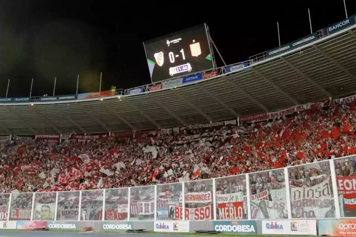 Soccer fans fill a stand with red and white flags while the stadium screen shows a 0-1 score during a night match. Soccer fans fill a stand with red and white flags while the stadium screen shows a 0-1 score during a night match.