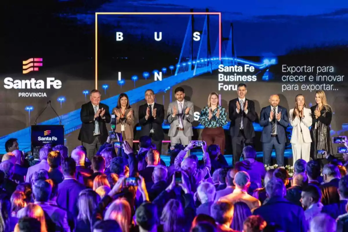 A group of people standing on a stage applauding in front of an audience at a business event in the province of Santa Fe, with screens in the background displaying the name of the forum and a lit-up bridge.