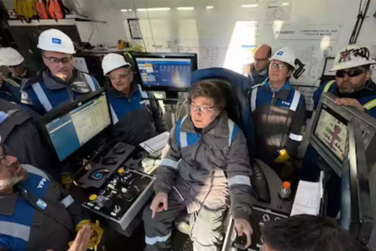 Varios trabajadores con cascos y uniformes de seguridad operan maquinaria y computadoras en una sala de control industrial