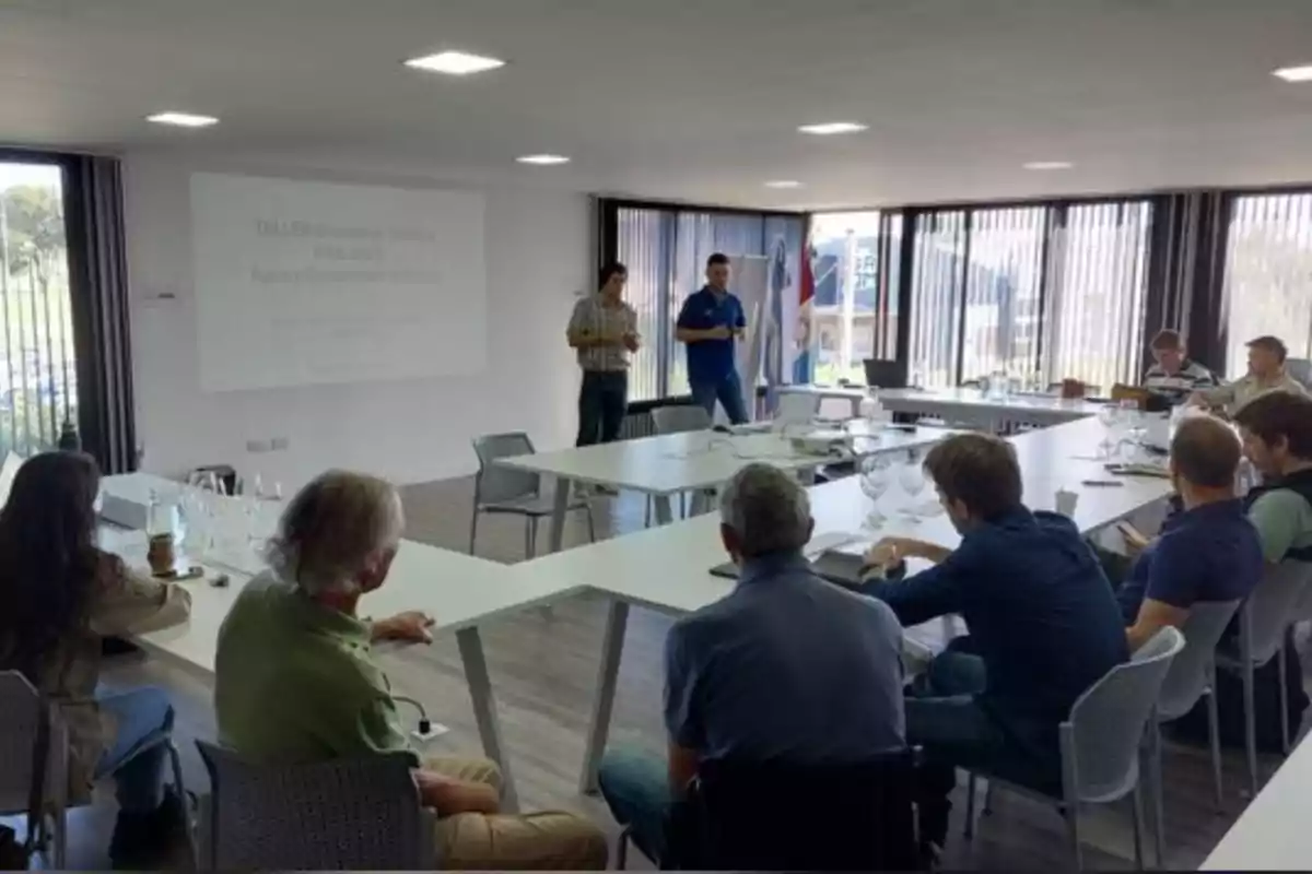 Personas sentadas alrededor de mesas en una sala de reuniones mientras dos hombres de pie presentan información proyectada en la pared Personas sentadas alrededor de mesas en una sala de reuniones mientras dos hombres de pie presentan información proyectada en la pared