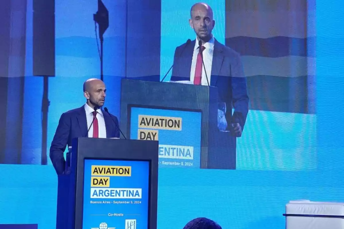 A man in a suit and red tie speaks at a podium during an event called Aviation Day Argentina, with a large screen behind him displaying his image.