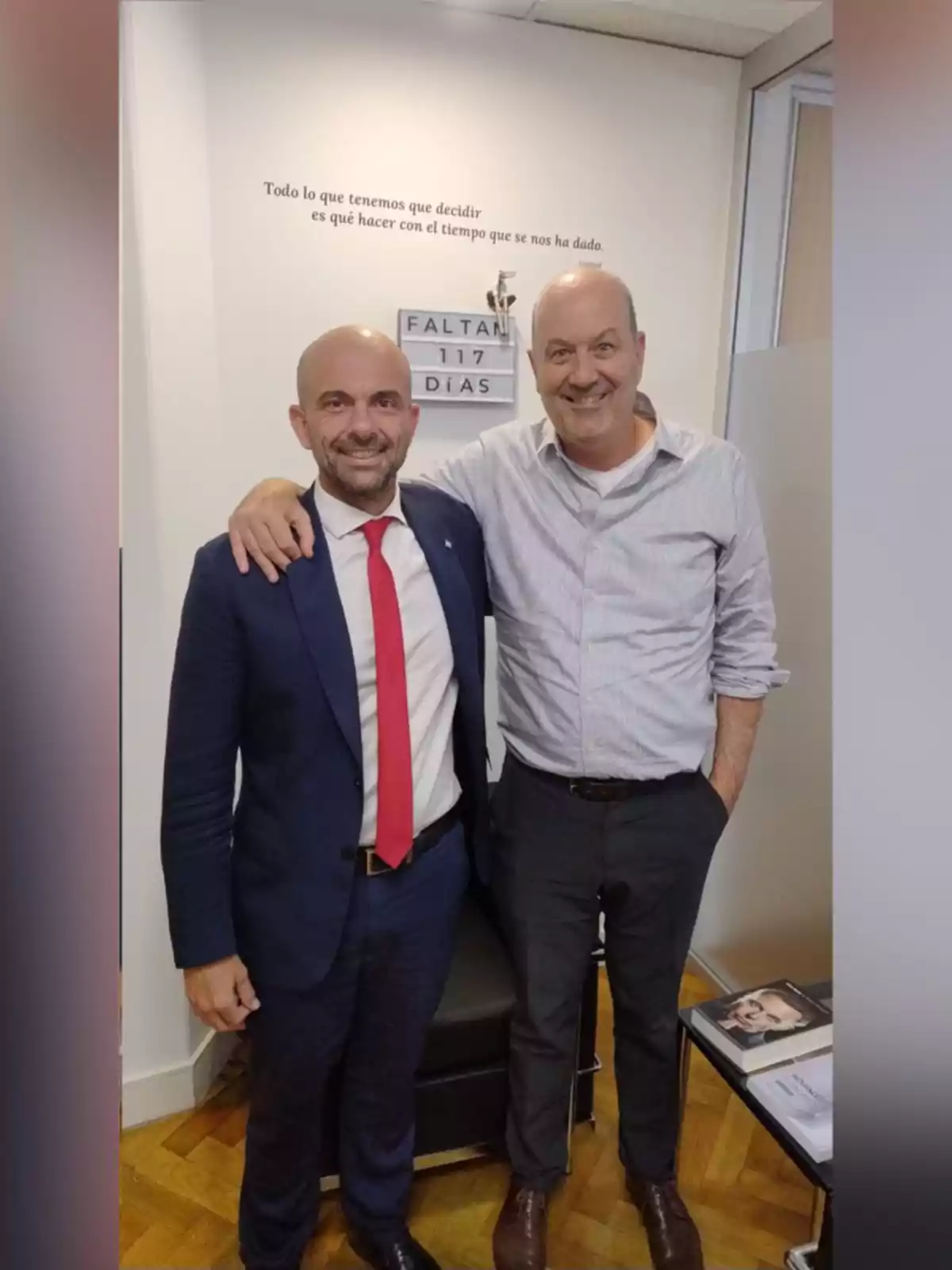 Dos hombres sonríen y posan juntos en una oficina elegante, uno de ellos lleva traje azul con corbata roja y el otro viste camisa clara y pantalón oscuro, al fondo hay una pared con una frase motivacional y un letrero que indica cuántos días faltan. Dos hombres sonríen y posan juntos en una oficina elegante, uno de ellos lleva traje azul con corbata roja y el otro viste camisa clara y pantalón oscuro, al fondo hay una pared con una frase motivacional y un letrero que indica cuántos días faltan.