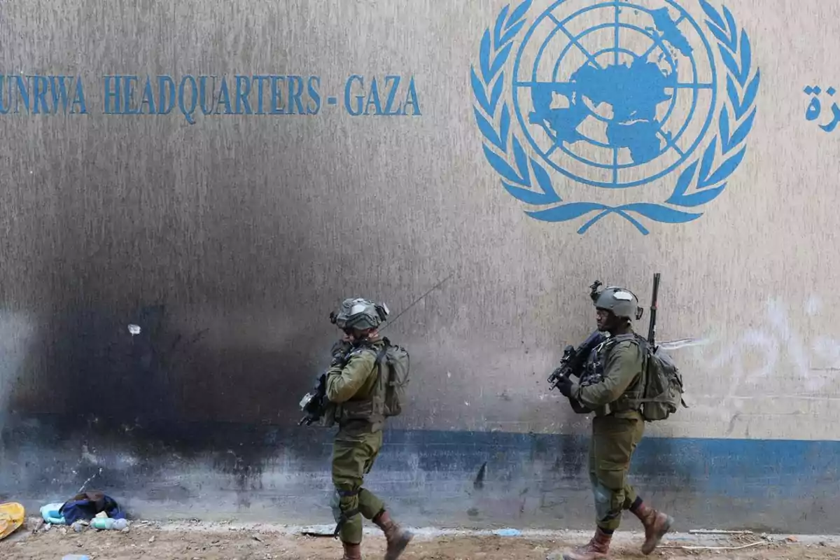 Dos soldados armados caminan frente a un muro con el logo de la ONU y la inscripción UNRWA Headquarters Gaza Dos soldados armados caminan frente a un muro con el logo de la ONU y la inscripción UNRWA Headquarters Gaza