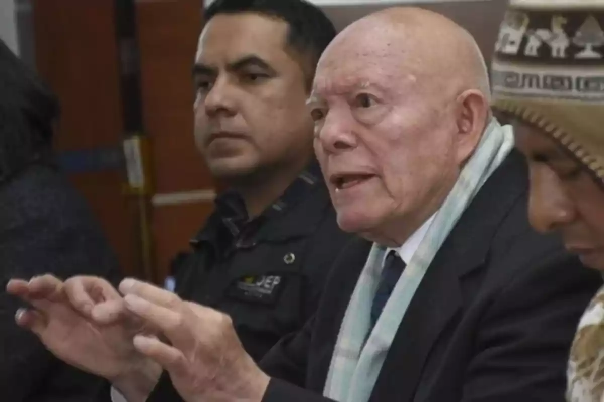 Un hombre mayor de cabello canoso y bufanda clara habla gesticulando con las manos mientras está sentado junto a otras personas en una reunión formal Un hombre mayor de cabello canoso y bufanda clara habla gesticulando con las manos mientras está sentado junto a otras personas en una reunión formal