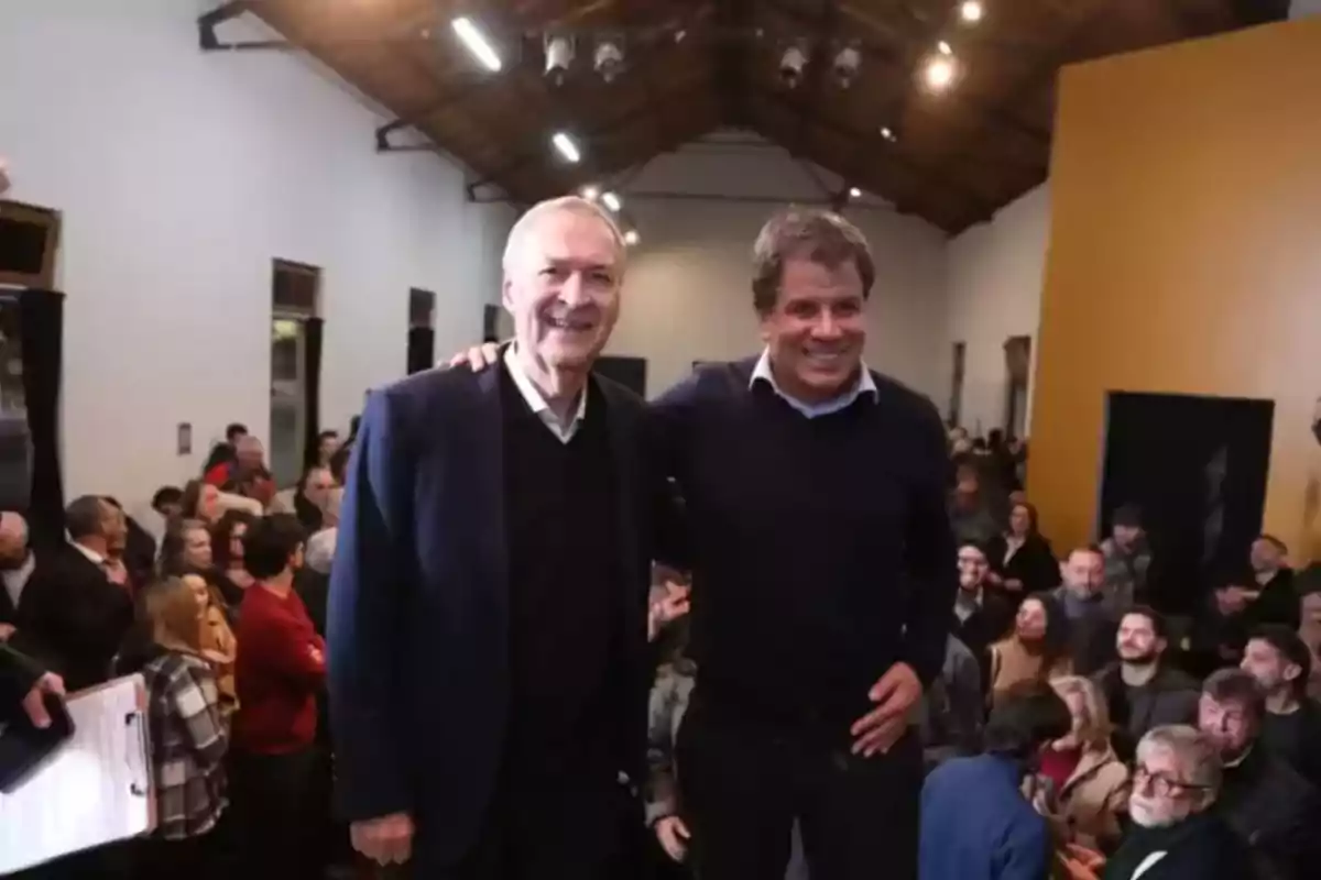 Dos hombres sonríen y posan juntos en el centro de una sala llena de personas, con un techo de madera y luces encendidas. Dos hombres sonríen y posan juntos en el centro de una sala llena de personas, con un techo de madera y luces encendidas.