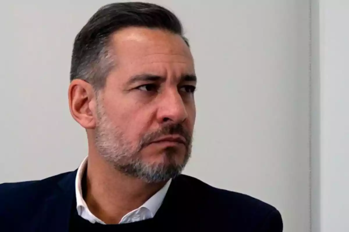 Hombre de cabello corto y barba con expresión seria mirando hacia un lado en un entorno interior. Hombre de cabello corto y barba con expresión seria mirando hacia un lado en un entorno interior.