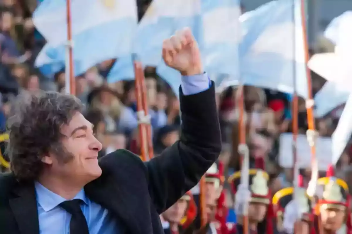 Hombre de cabello rizado con traje oscuro levantando el puño en alto mientras sonríe, con banderas de Argentina y personas uniformadas al fondo Hombre de cabello rizado con traje oscuro levantando el puño en alto mientras sonríe, con banderas de Argentina y personas uniformadas al fondo