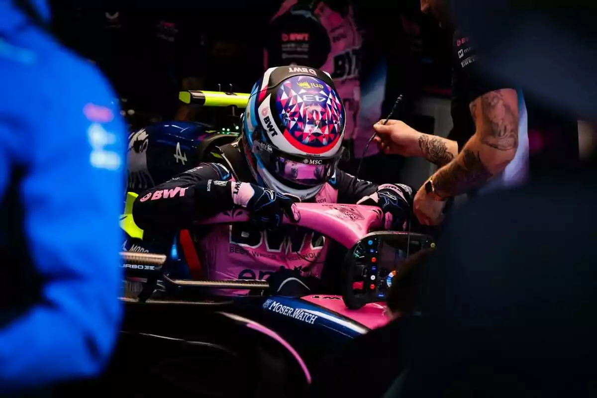 Piloto de Fórmula 1 con casco y uniforme rosa y azul sentado en su monoplaza dentro del garaje mientras un mecánico lo asiste