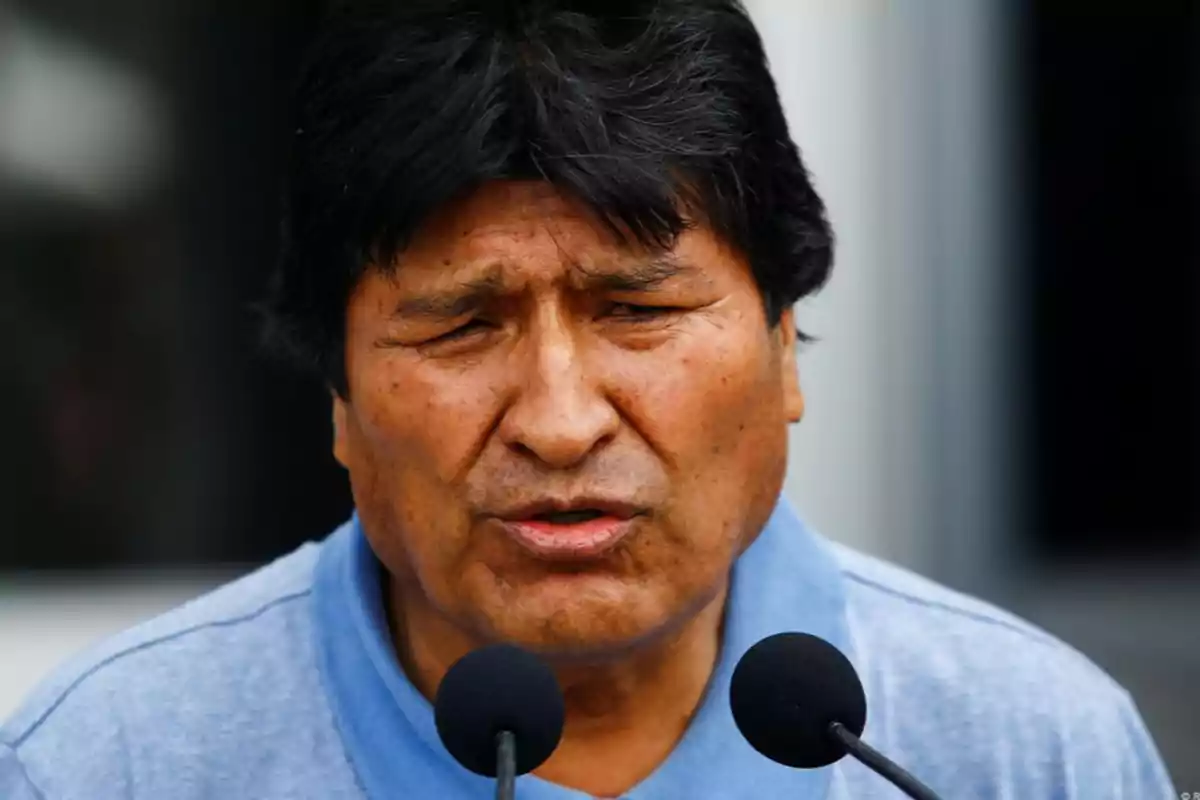 Dark-haired man speaking in front of two microphones with a serious expression, wearing a blue t-jersey