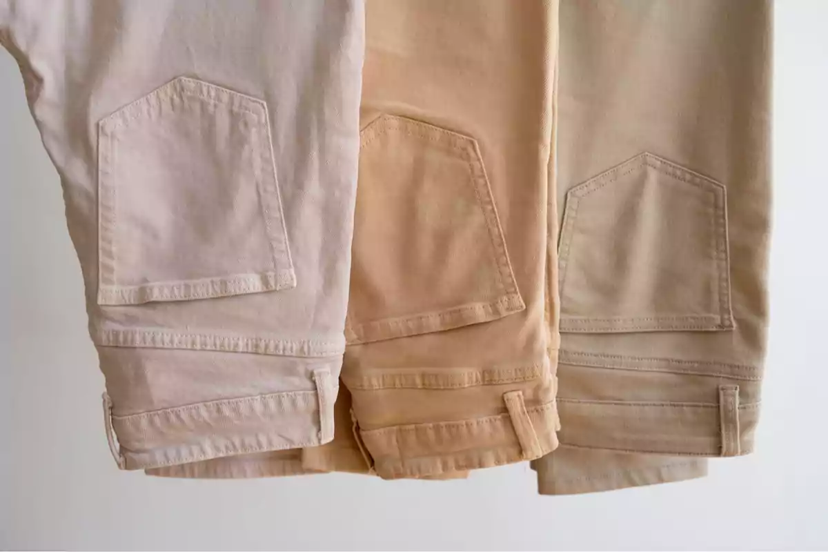 Three beige-toned denim pants hanging side by side, showing the back pockets. Three beige-toned denim pants hanging side by side, showing the back pockets.