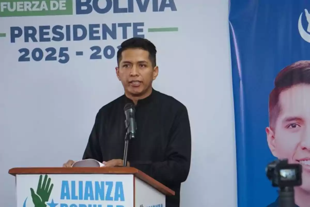 A man with short dark hair speaks into a microphone at a podium with the Alianza Popular logo; behind him, there is a presidential campaign poster for Bolivia 2025-2030. A man with short dark hair speaks into a microphone at a podium with the Alianza Popular logo; behind him, there is a presidential campaign poster for Bolivia 2025-2030.