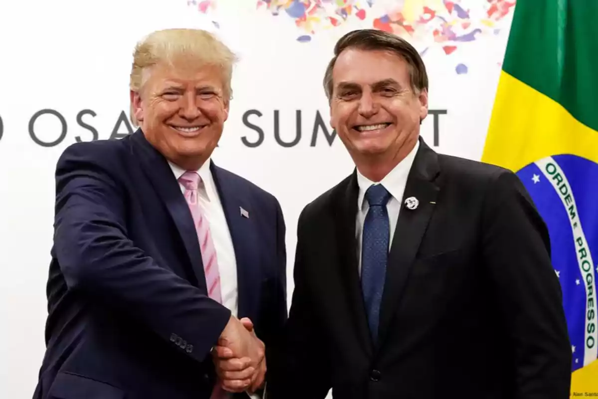 Dos hombres de traje sonríen y se dan la mano frente a una bandera de Brasil y un fondo con letras y colores.