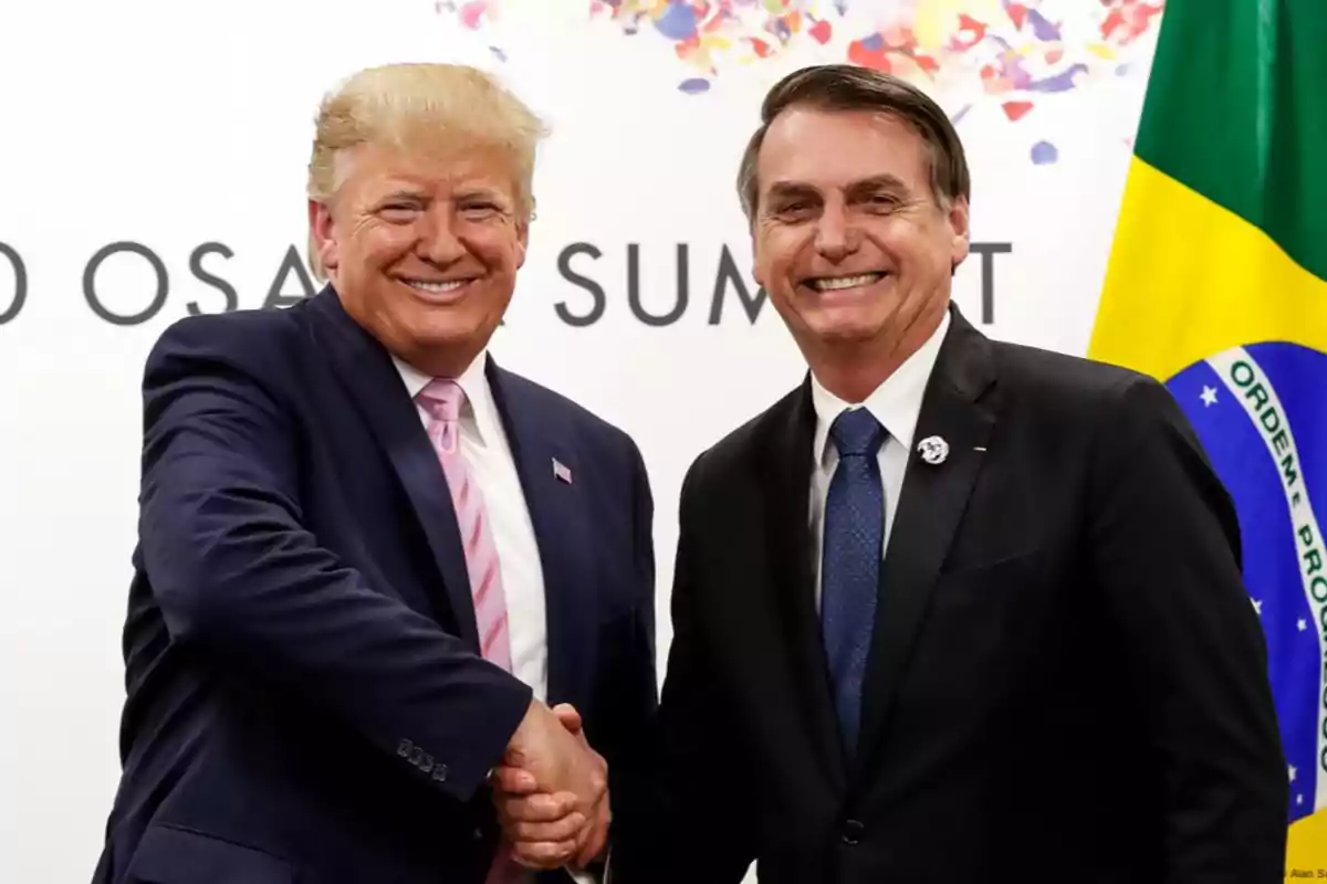 Two men in suits smile and shake hands in front of a Brazilian flag and a background with festive letters and colors.