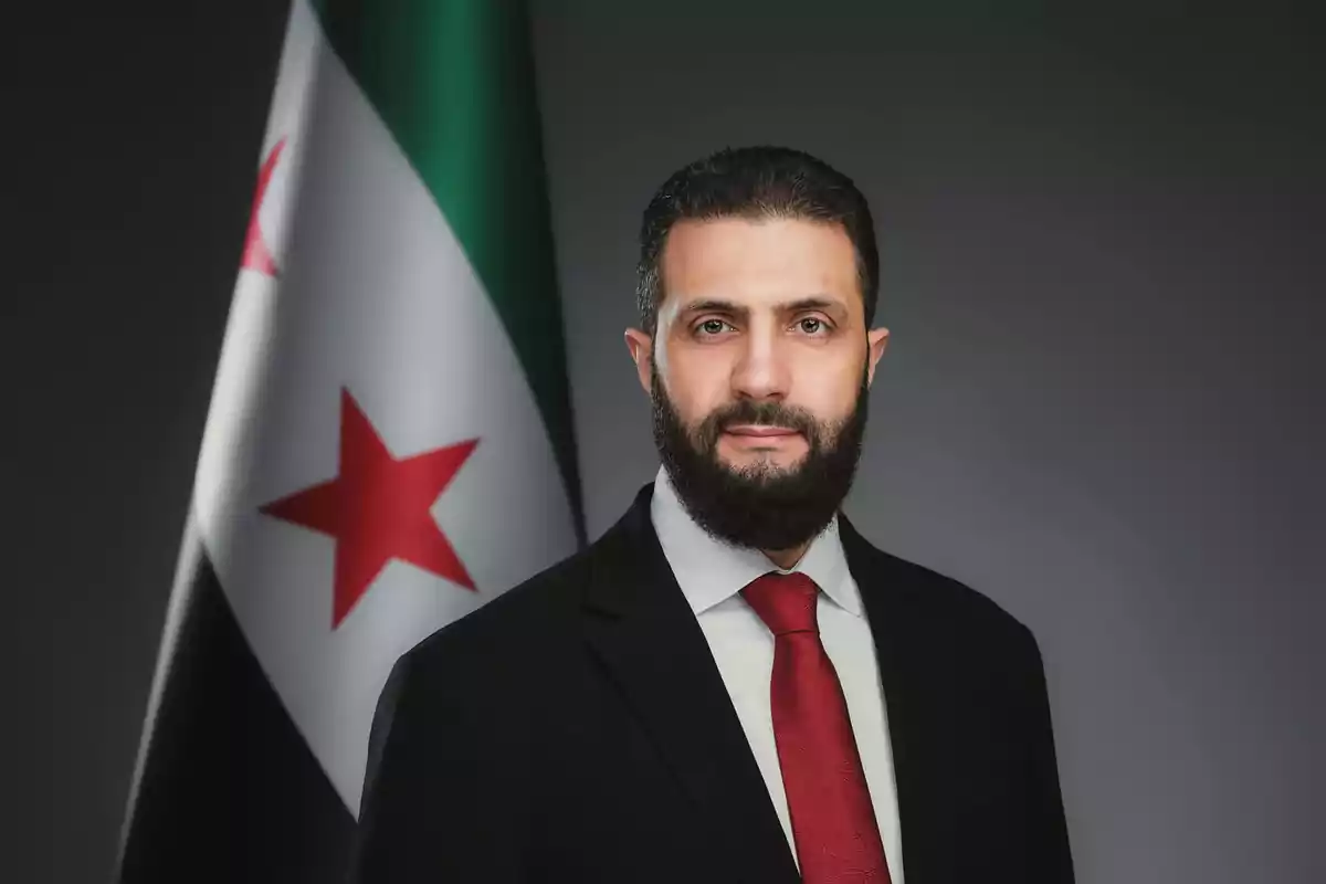 Man with a beard and a dark suit with a red tie posing in front of a flag with red stars and green, white, and black stripes against a gray background.