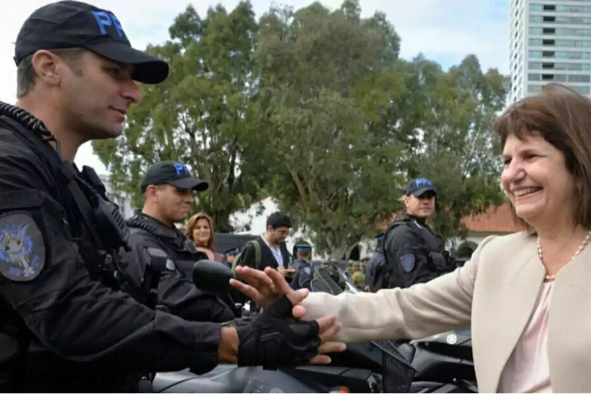 Una mujer sonriente saluda a un policía uniformado mientras otros agentes observan en un entorno al aire libre con árboles y edificios de fondo Una mujer sonriente saluda a un policía uniformado mientras otros agentes observan en un entorno al aire libre con árboles y edificios de fondo