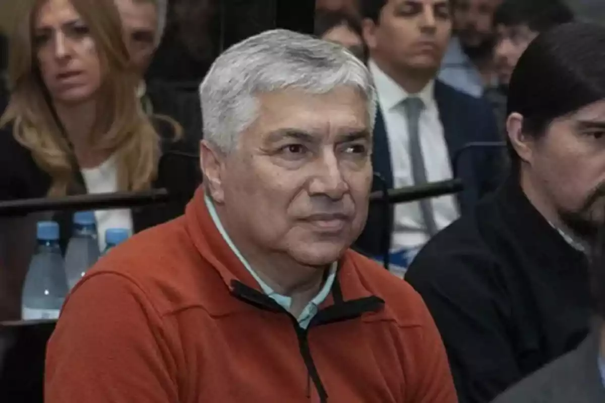 Gray-haired man in an orange sweater sitting among other people in a formal setting