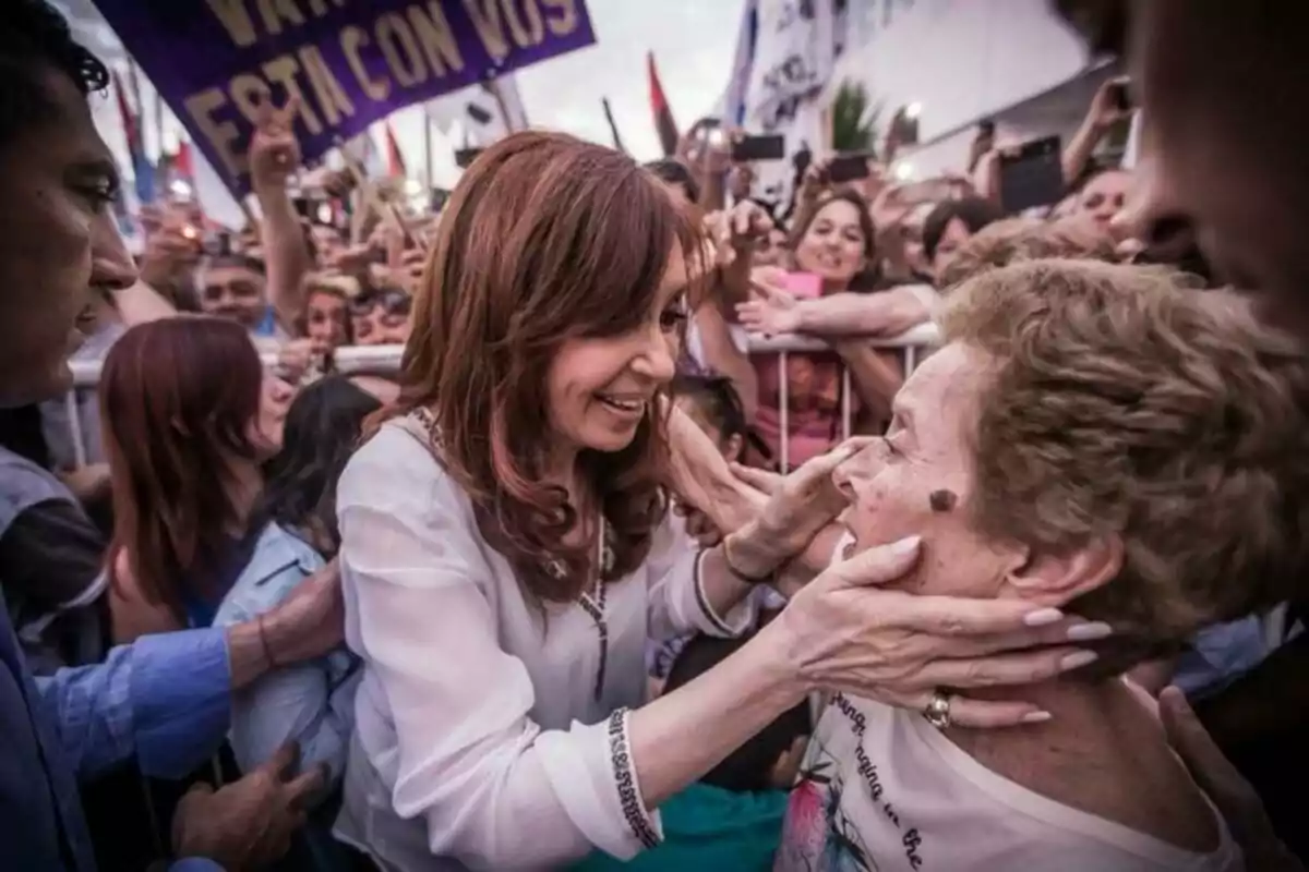 A smiling woman holds the face of an elderly lady while a crowd watches and takes photos at an outdoor event.