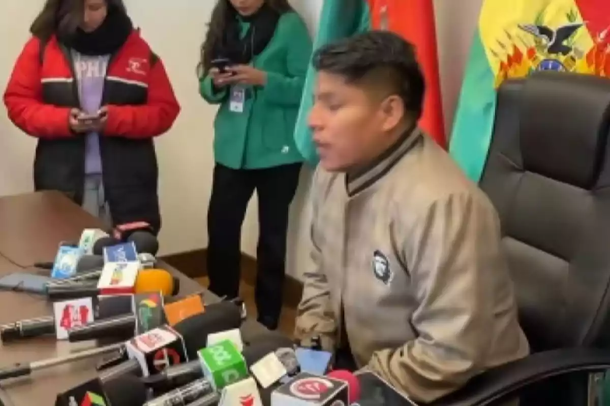 A man sitting in a chair in front of several microphones during a press conference, with two people standing in the background and Bolivian flags behind them. A man sitting in a chair in front of several microphones during a press conference, with two people standing in the background and Bolivian flags behind them.