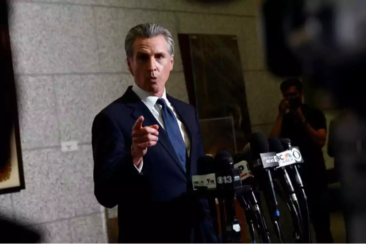 Man in a dark blue suit speaking in front of several microphones at a press conference inside a building