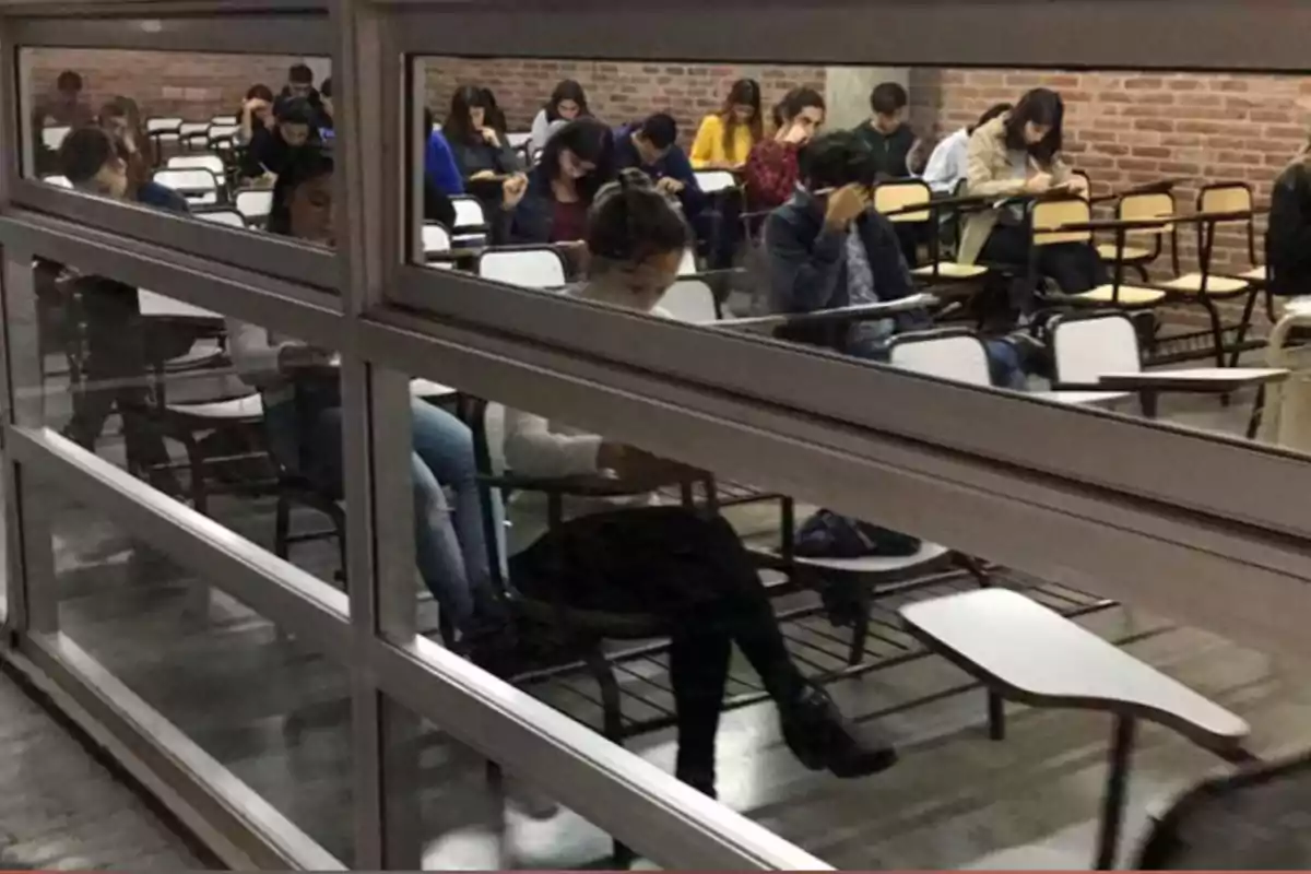 Varios estudiantes sentados en un salón de clases tomando un examen, vistos a través de una ventana interior Varios estudiantes sentados en un salón de clases tomando un examen, vistos a través de una ventana interior