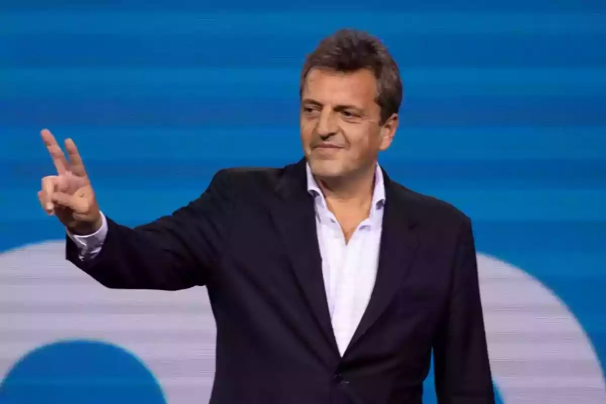 Hombre de traje oscuro haciendo el gesto de la victoria con los dedos frente a un fondo azul Hombre de traje oscuro haciendo el gesto de la victoria con los dedos frente a un fondo azul