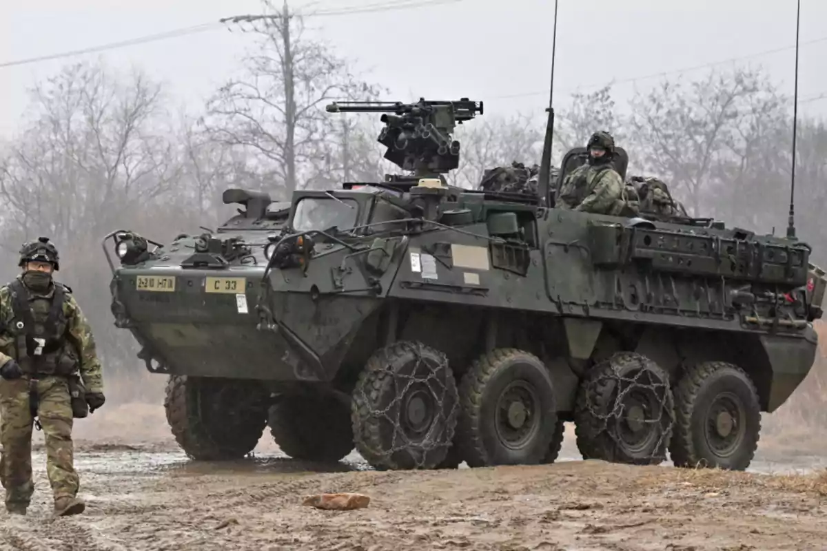 Vehículo militar blindado de ocho ruedas con soldados a bordo y un soldado caminando al lado en un entorno boscoso y fangoso Vehículo militar blindado de ocho ruedas con soldados a bordo y un soldado caminando al lado en un entorno boscoso y fangoso