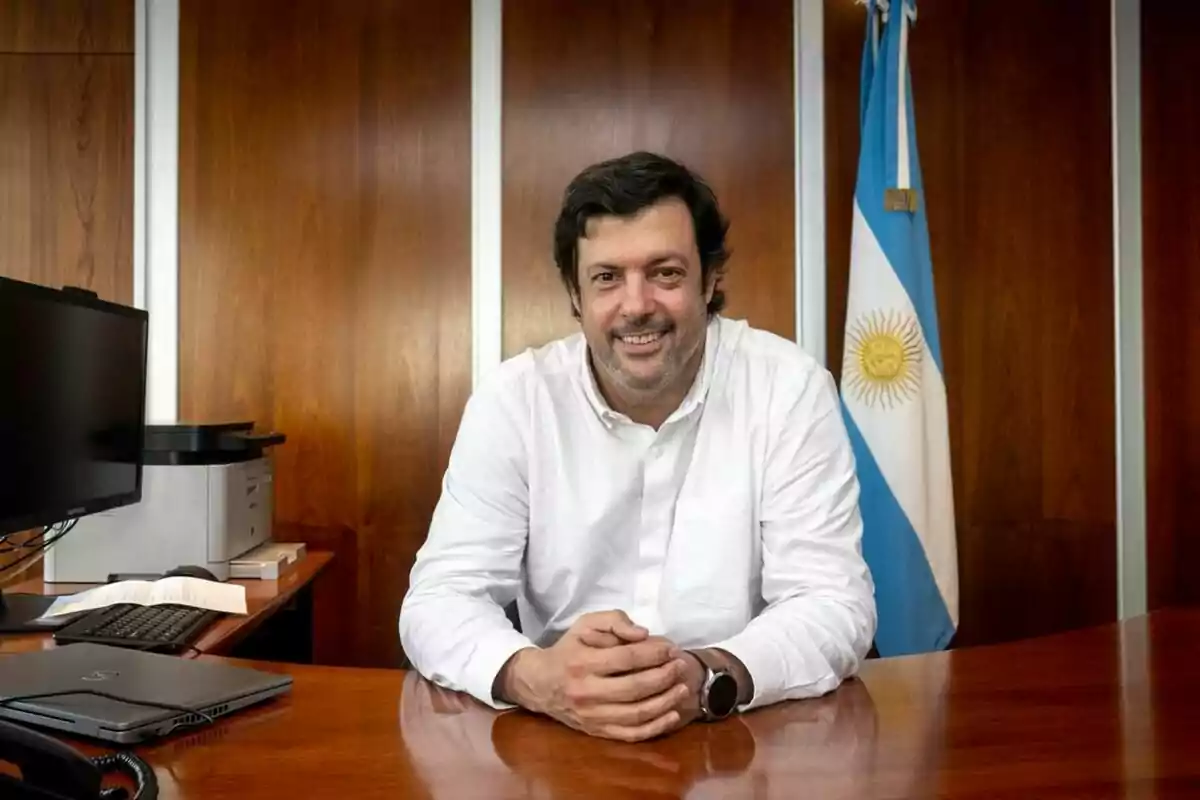 Hombre de cabello oscuro y barba sentado en un escritorio de oficina con una bandera de Argentina al fondo Hombre de cabello oscuro y barba sentado en un escritorio de oficina con una bandera de Argentina al fondo