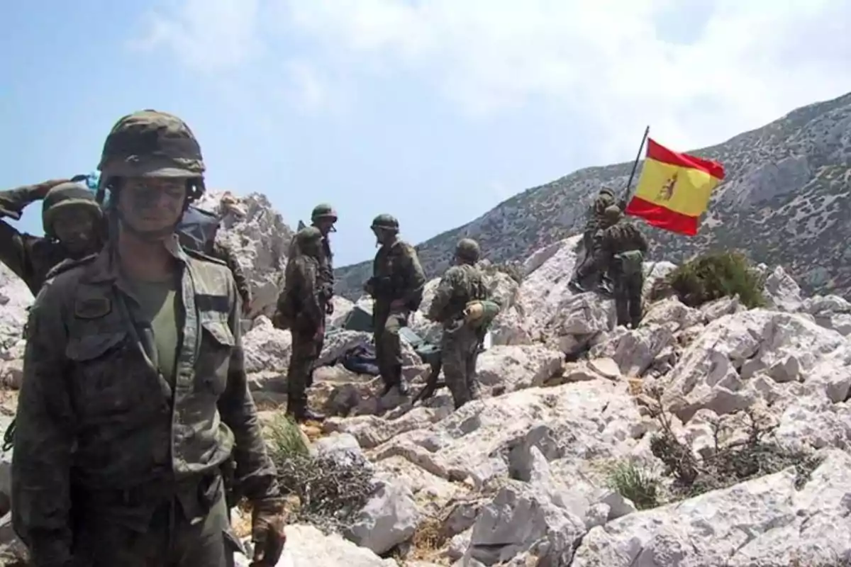 Soldados con uniforme de camuflaje en una zona rocosa mientras uno de ellos sostiene una bandera de España Soldados con uniforme de camuflaje en una zona rocosa mientras uno de ellos sostiene una bandera de España