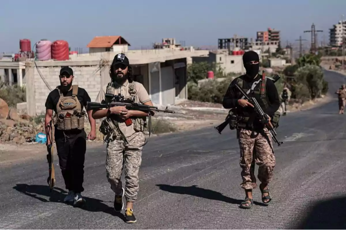 Tres hombres armados con equipo militar caminan por una carretera en una zona urbana con edificios en construcción al fondo Tres hombres armados con equipo militar caminan por una carretera en una zona urbana con edificios en construcción al fondo