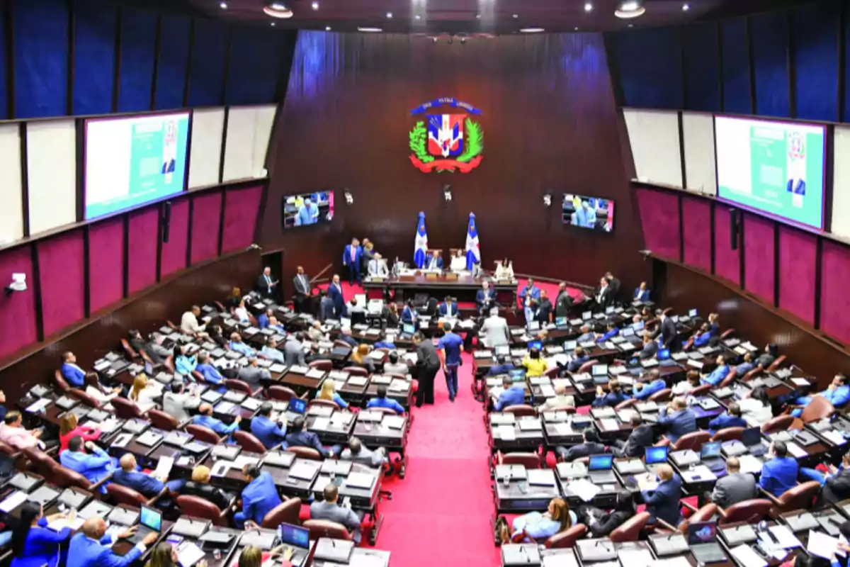 Vista general del salón de sesiones de la Cámara de Diputados de la República Dominicana con legisladores sentados en sus escritorios y varias personas de pie en el centro