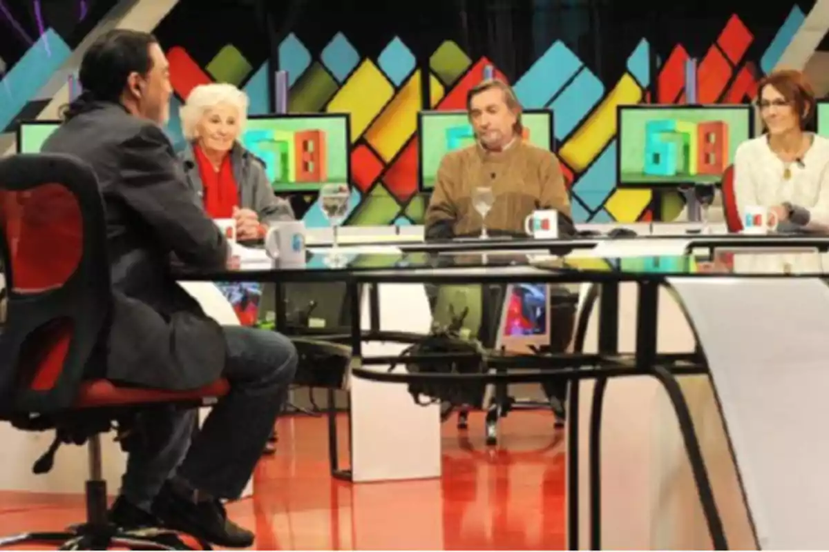 Cuatro personas sentadas alrededor de una mesa en un estudio de televisión con pantallas y fondo colorido Cuatro personas sentadas alrededor de una mesa en un estudio de televisión con pantallas y fondo colorido