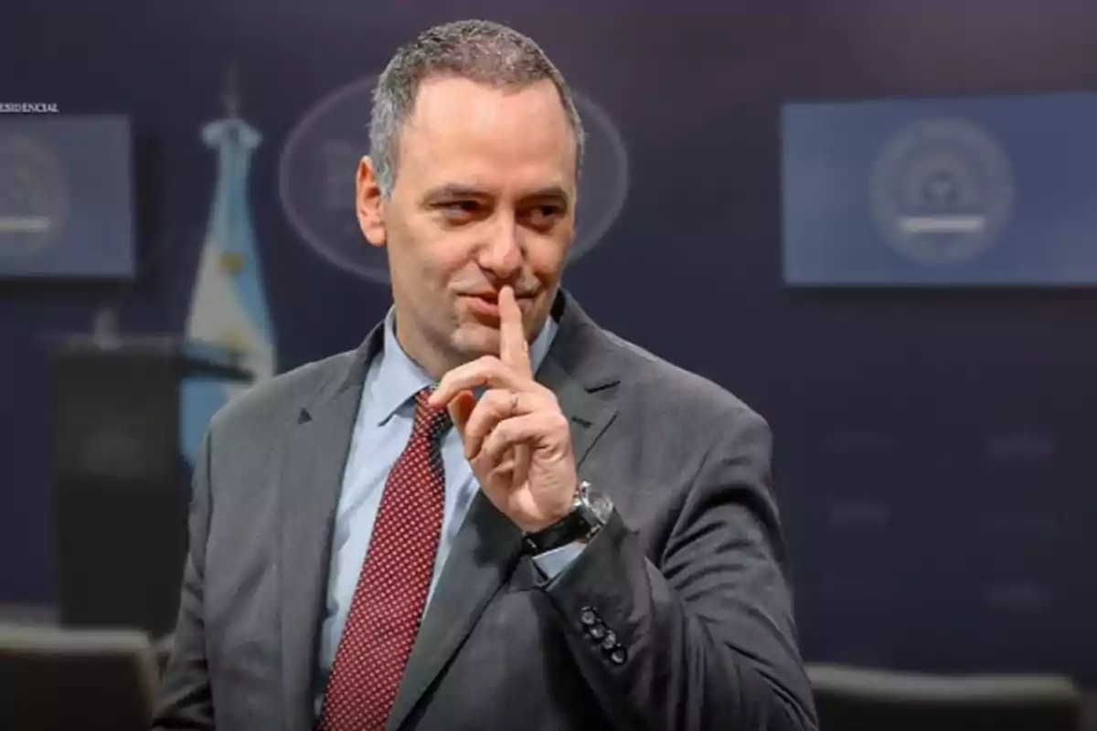 Hombre de traje y corbata haciendo el gesto de silencio con el dedo índice frente a los labios en un entorno institucional. Hombre de traje y corbata haciendo el gesto de silencio con el dedo índice frente a los labios en un entorno institucional.