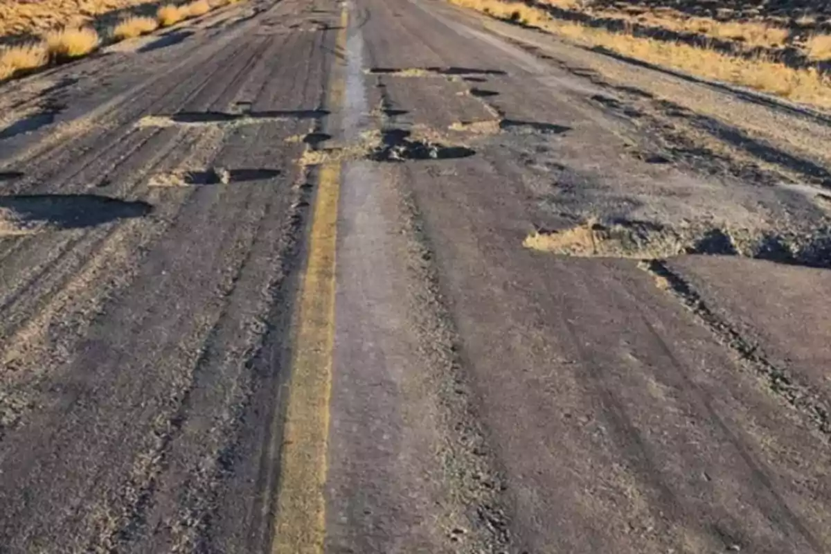 Road with numerous potholes and worn asphalt surrounded by dry vegetation. Road with numerous potholes and worn asphalt surrounded by dry vegetation.