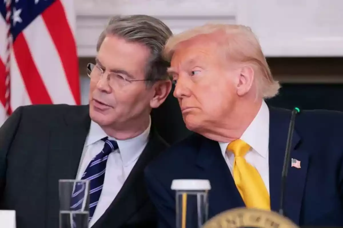 Dos hombres de traje conversan en una reunión oficial con una bandera de Estados Unidos al fondo