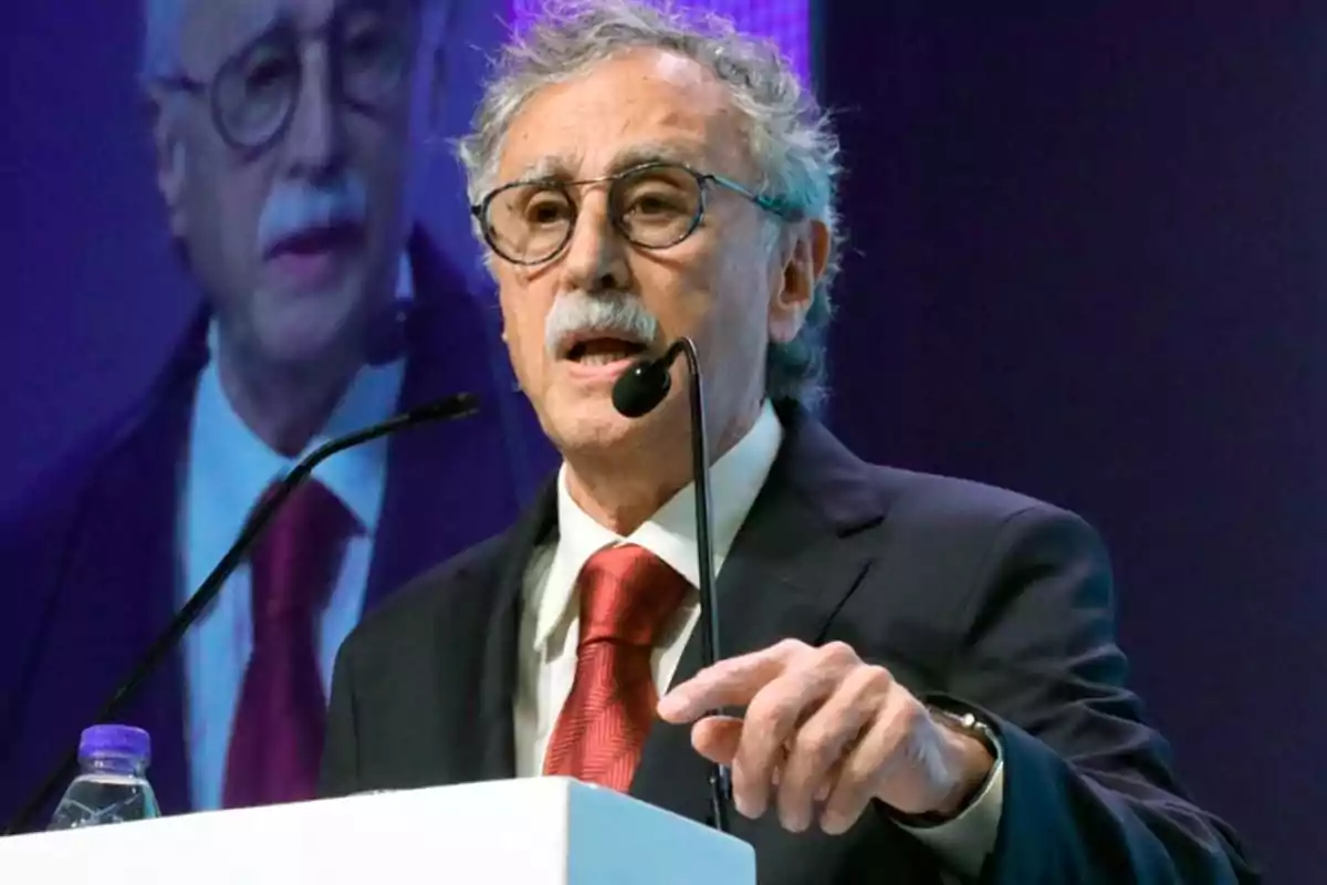 Gray-haired man with glasses speaking at a podium with a microphone, wearing a dark suit and red tie, with his image projected on a screen behind him Gray-haired man with glasses speaking at a podium with a microphone, wearing a dark suit and red tie, with his image projected on a screen behind him