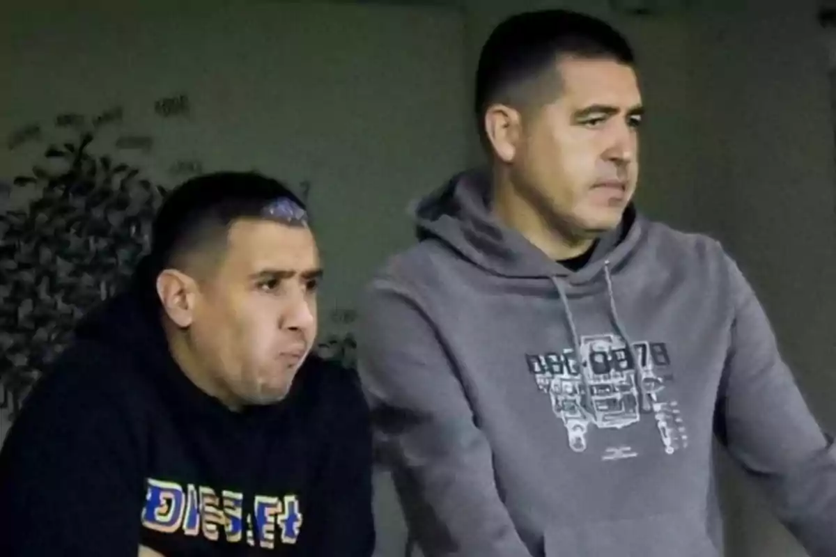 Dos hombres con expresión seria observan algo fuera de la imagen uno lleva una sudadera negra y el otro una sudadera gris