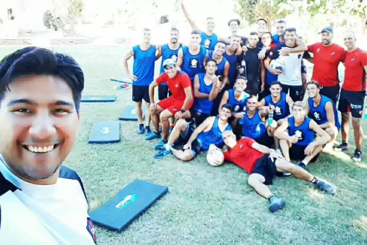A group of young athletes smiles and poses for a selfie outdoors on a grassy field, some wearing blue jerseys and others red, while in the foreground a man is smiling at the camera. A group of young athletes smiles and poses for a selfie outdoors on a grassy field, some wearing blue jerseys and others red, while in the foreground a man is smiling at the camera.