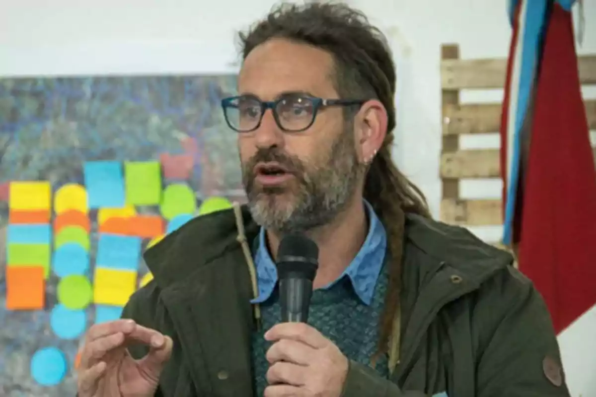 Man with glasses and a beard speaking while holding a microphone, with colorful sticky notes and a flag in the background. Man with glasses and a beard speaking while holding a microphone, with colorful sticky notes and a flag in the background.
