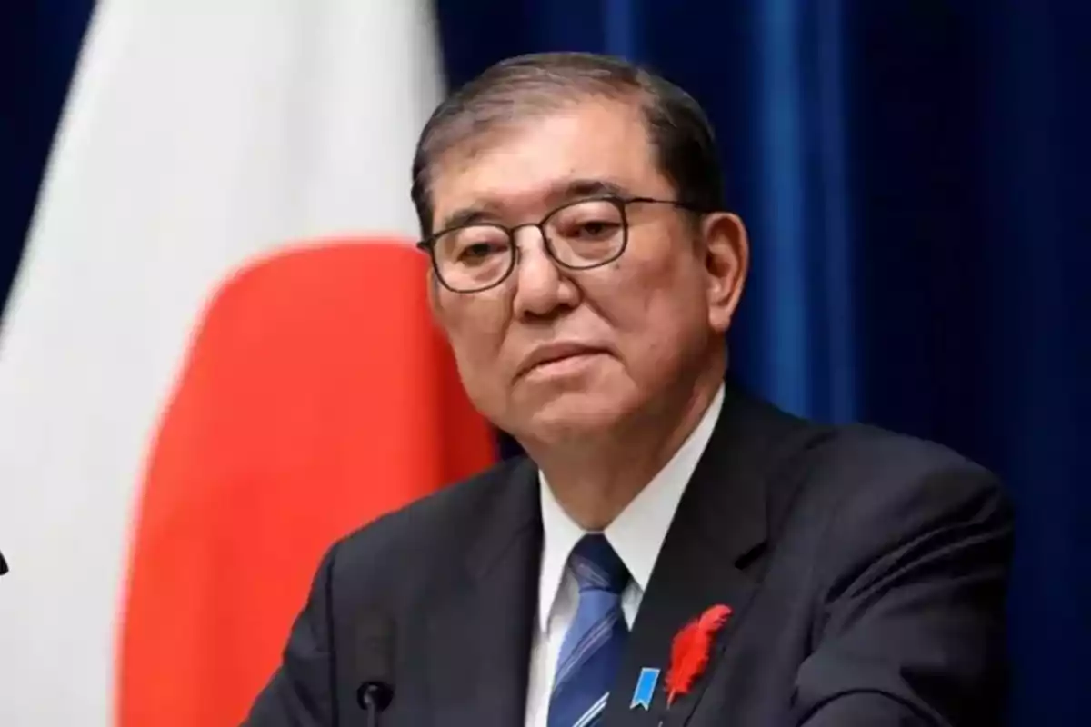 Hombre de traje y gafas con expresión seria frente a una bandera de Japón