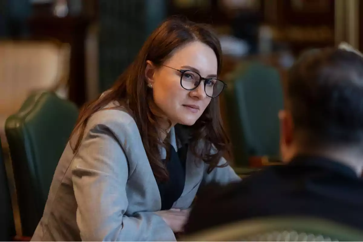 Mujer con gafas y saco gris sentada en una reunión mirando atentamente a otra persona Mujer con gafas y saco gris sentada en una reunión mirando atentamente a otra persona