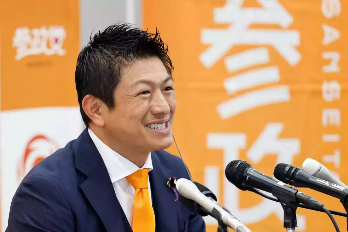 Hombre sonriente con traje azul y corbata naranja hablando frente a varios micrófonos con un fondo naranja y blanco con letras japonesas. Hombre sonriente con traje azul y corbata naranja hablando frente a varios micrófonos con un fondo naranja y blanco con letras japonesas.