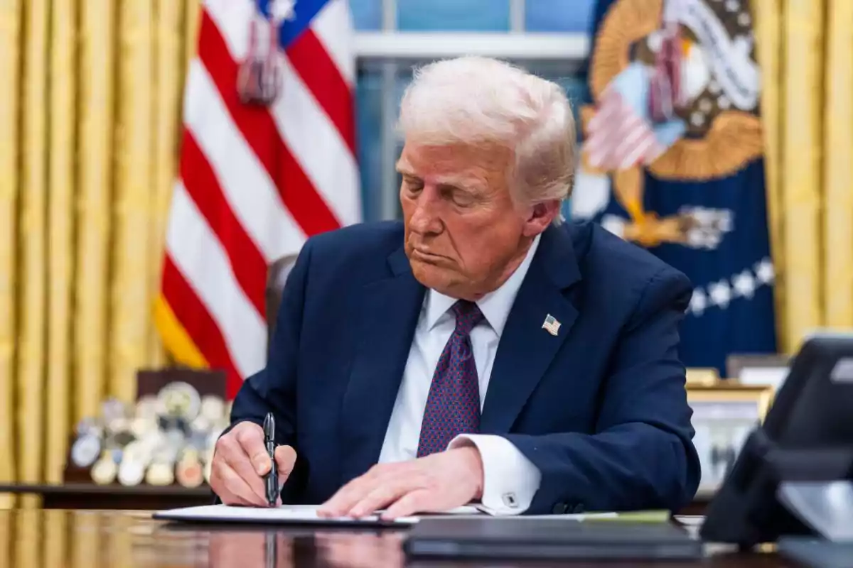 Blond-haired man in a blue suit signing a document in an official office with a United States flag in the background