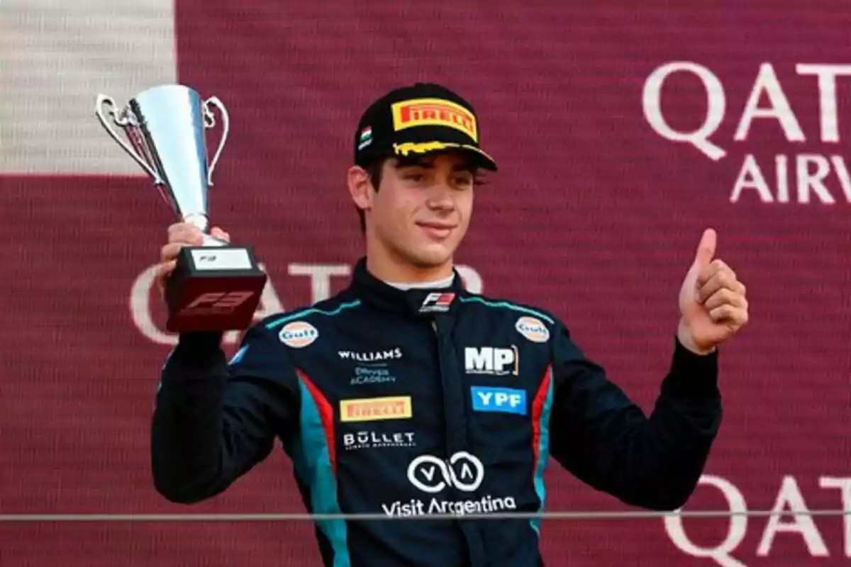 Piloto de automovilismo celebrando en el podio con un trofeo en una mano y haciendo el gesto de pulgar arriba con la otra mano Piloto de automovilismo celebrando en el podio con un trofeo en una mano y haciendo el gesto de pulgar arriba con la otra mano
