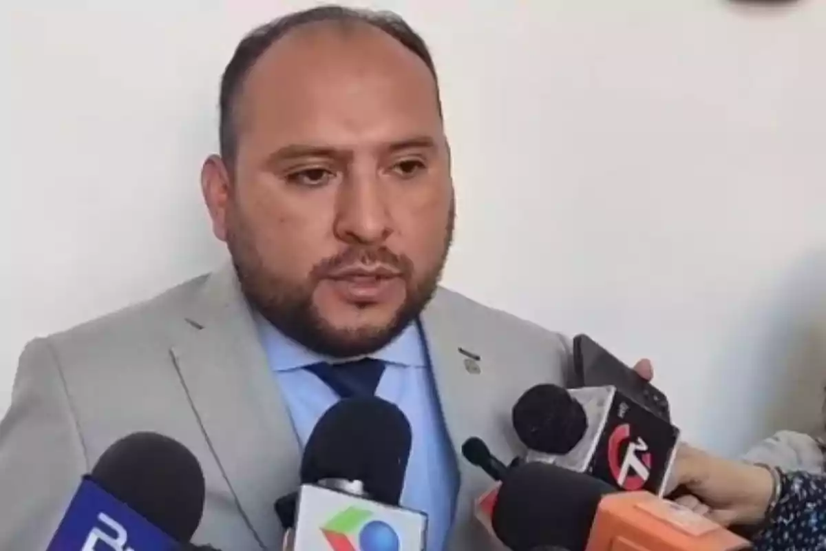 Hombre con saco gris y camisa azul hablando frente a varios micrófonos de diferentes medios de comunicación. Hombre con saco gris y camisa azul hablando frente a varios micrófonos de diferentes medios de comunicación.