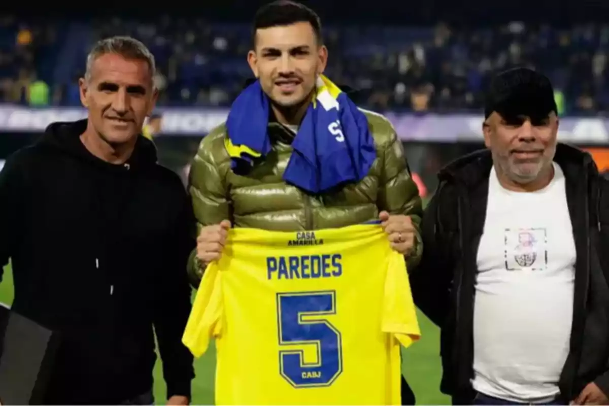 Three people pose in a soccer stadium while one of them holds a yellow jersey with the name Paredes and the number 5. Three people pose in a soccer stadium while one of them holds a yellow jersey with the name Paredes and the number 5.