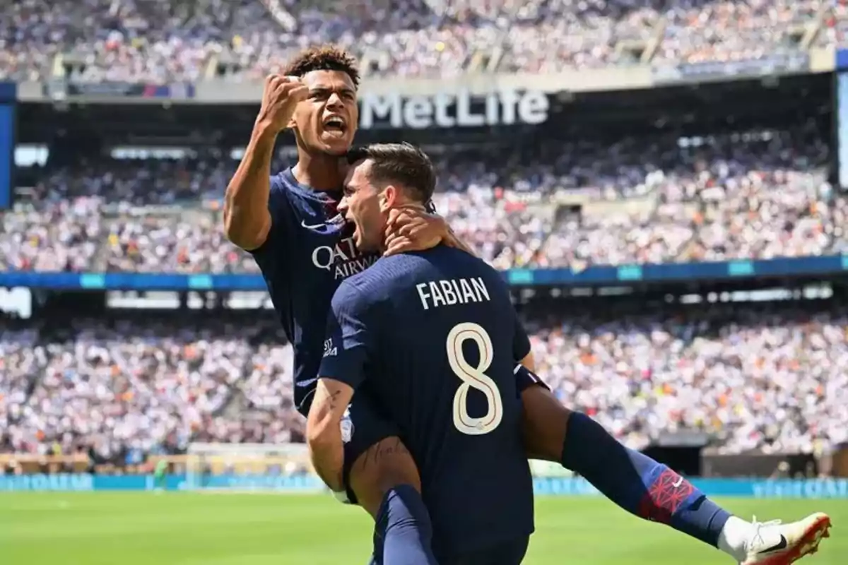 Two players celebrate a goal in a packed stadium; one of them raises his fist while the other holds him in his arms and wears the name Fabián and the number 8 on his jersey. Two players celebrate a goal in a packed stadium; one of them raises his fist while the other holds him in his arms and wears the name Fabián and the number 8 on his jersey.