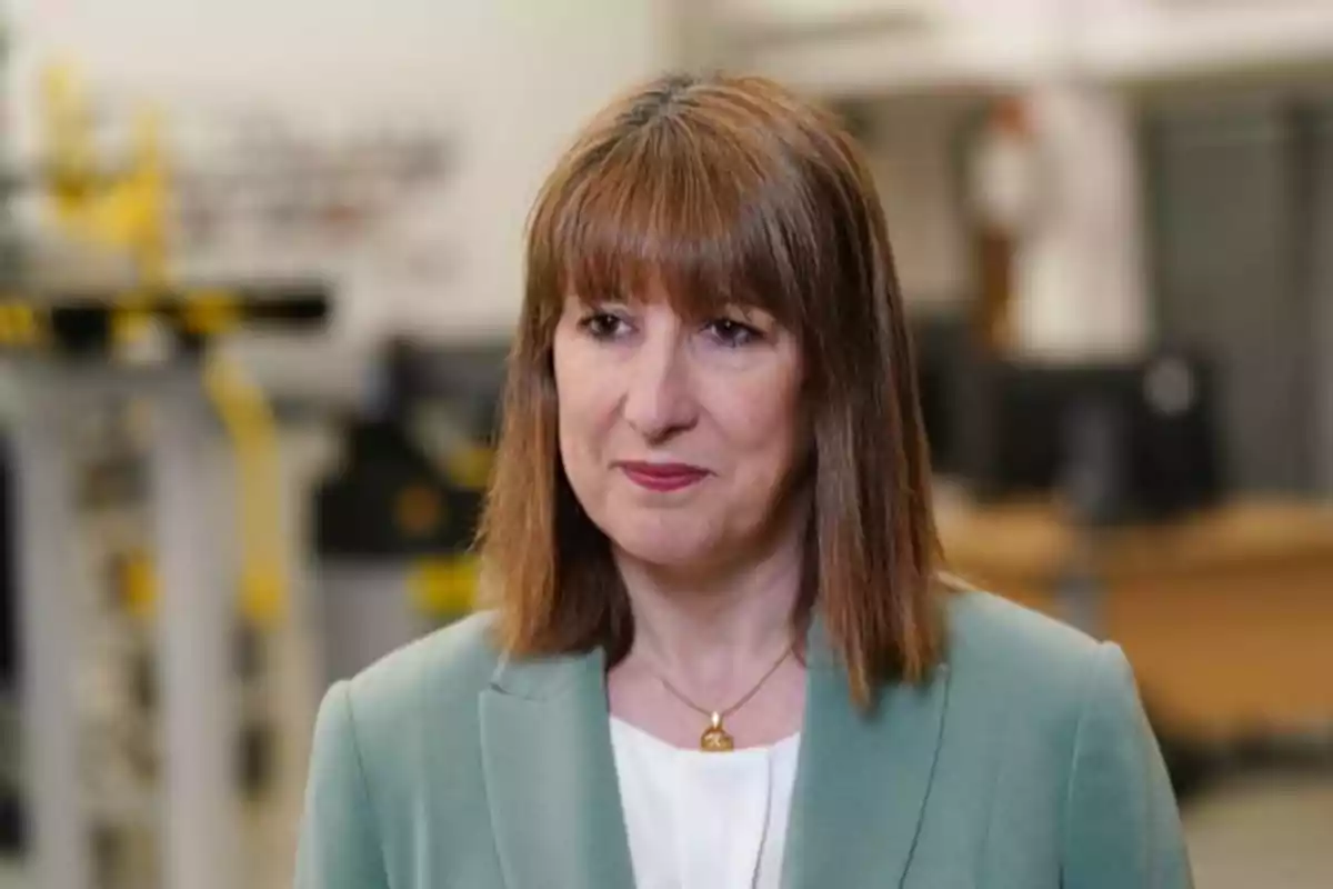Brown-haired woman with straight hair and bangs, wearing a light green blazer and a white blouse, in a blurred office setting. Brown-haired woman with straight hair and bangs, wearing a light green blazer and a white blouse, in a blurred office setting.